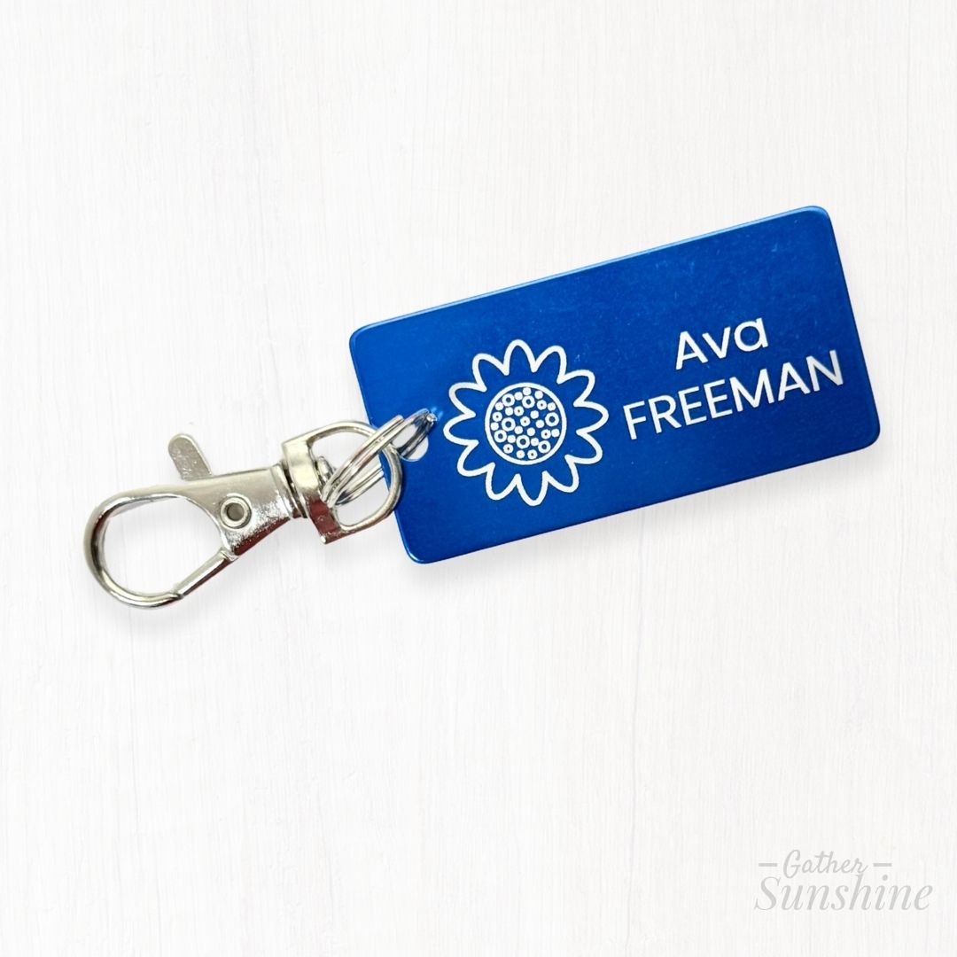 Close-up of blue 1x2 inch kids tag with engraved flower design, lobster clasp, and custom name engraving