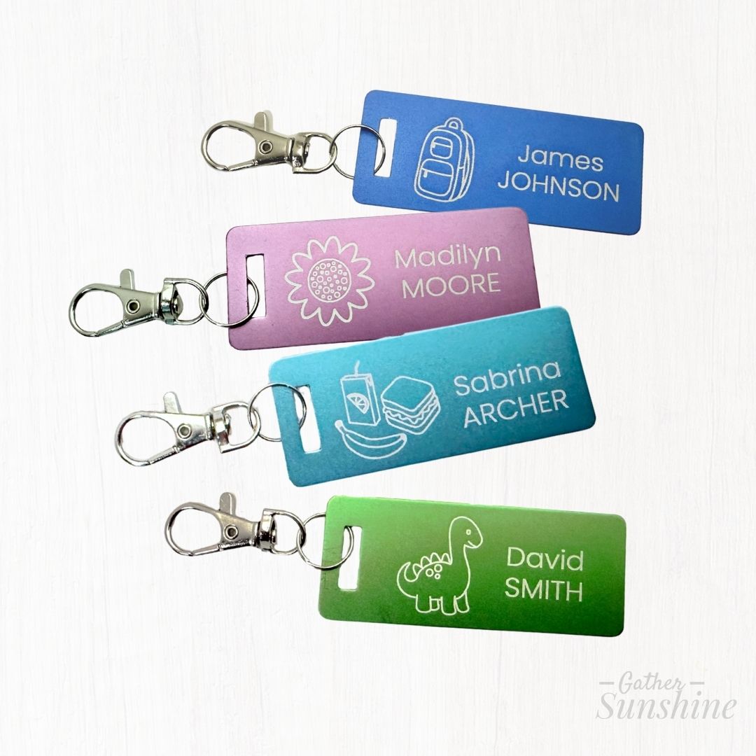Set of four colorful 1x3 inch kids luggage tags, each with a lobster clasp: a purple flower tag, blue backpack tag, green dinosaur tag, and teal lunch tag — all personalized with names. Perfect for backpacks, lunch boxes, and more. GatherSunshine.