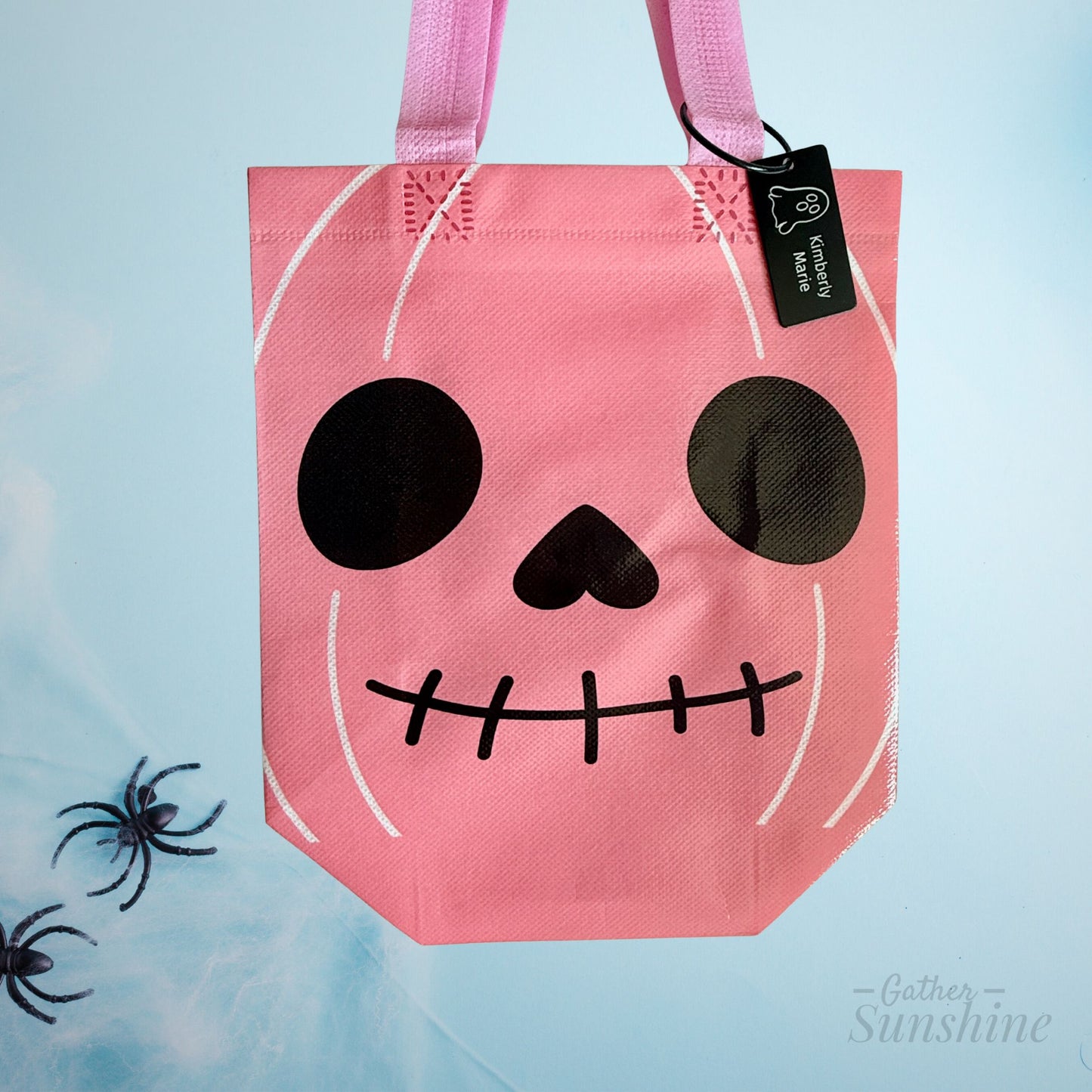 Pink Halloween-themed tote bag with a skull design on a light blue background. The bag has a custom engrave ghost tag made by Gather Sunshine.
