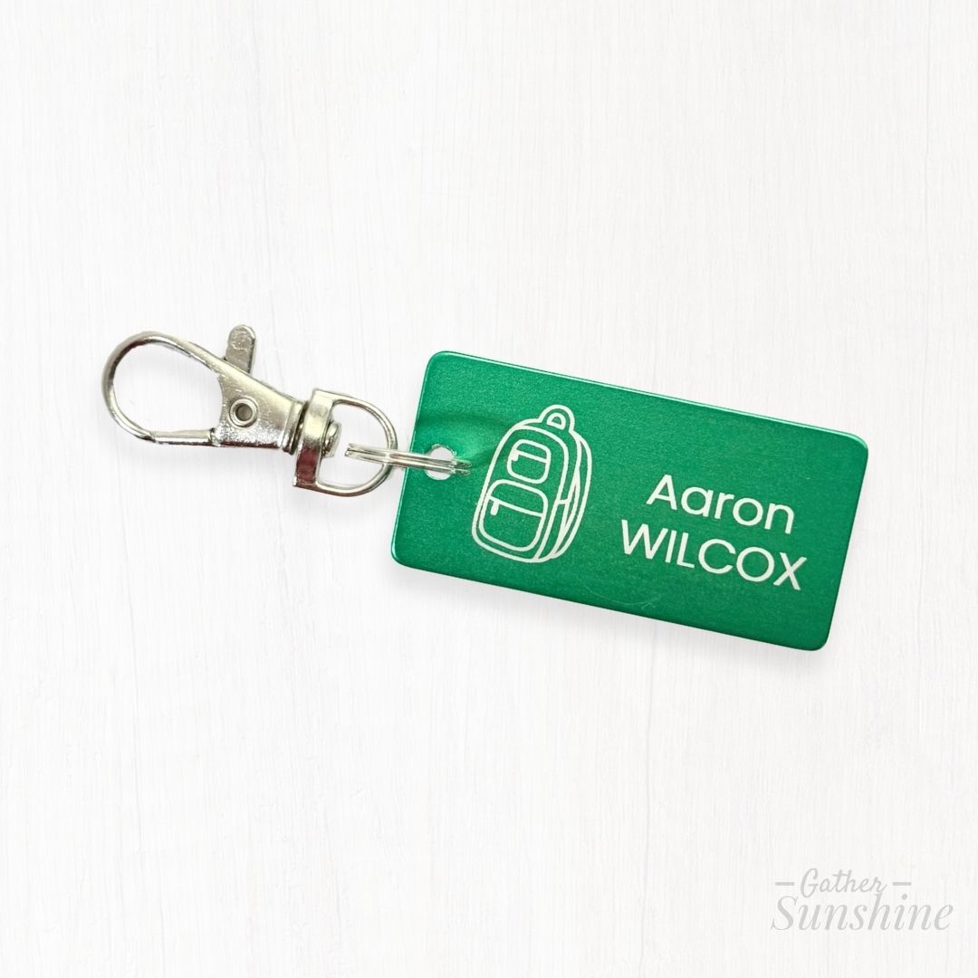 Close-up of green 1x2 inch kids tag with engraved backpack icon, lobster clasp, and personalized name engraving