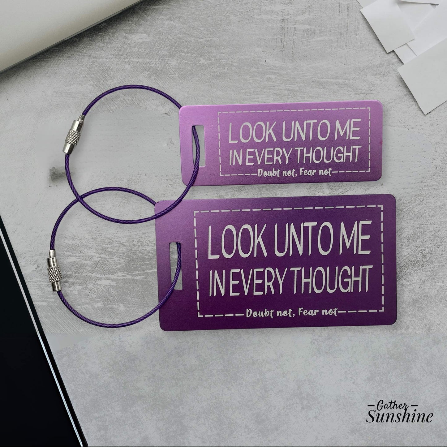 Small (3"x1") and large (3.5"x2") purple luggage tags with motivational text "Look Unto Me In Every Thought, Doubt Not, Fear Not" on a gray surface. Engraved by Gather Sunshine