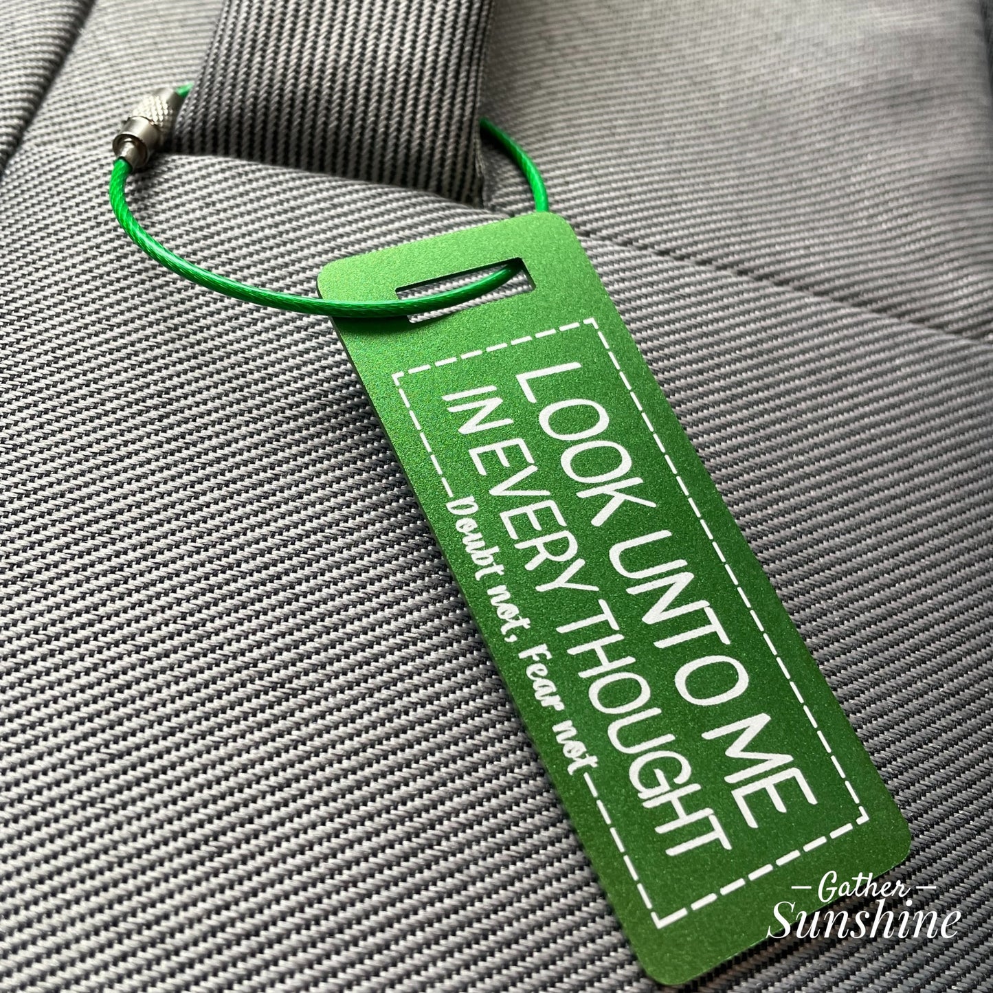 Small (3"x1") green luggage tag with motivational text "Look Unto Me In Every Thought, Doubt Not, Fear Not" attached to gray suitcase by Gather Sunshine.