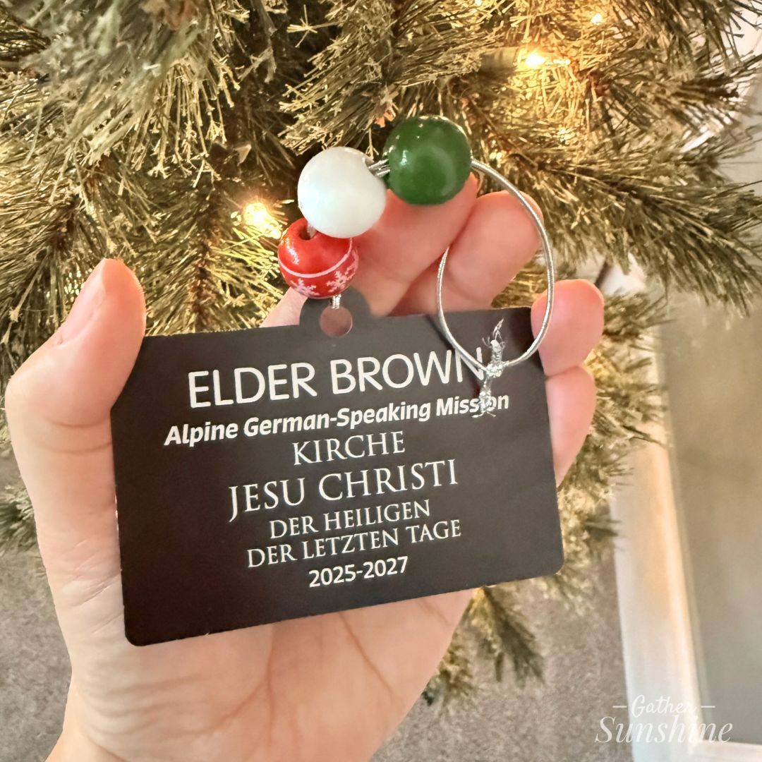 LDS Missionary Tag Christmas ornament in German hanging in a hand next to a decorated tree. Custome engraved by Gather Sunshine.