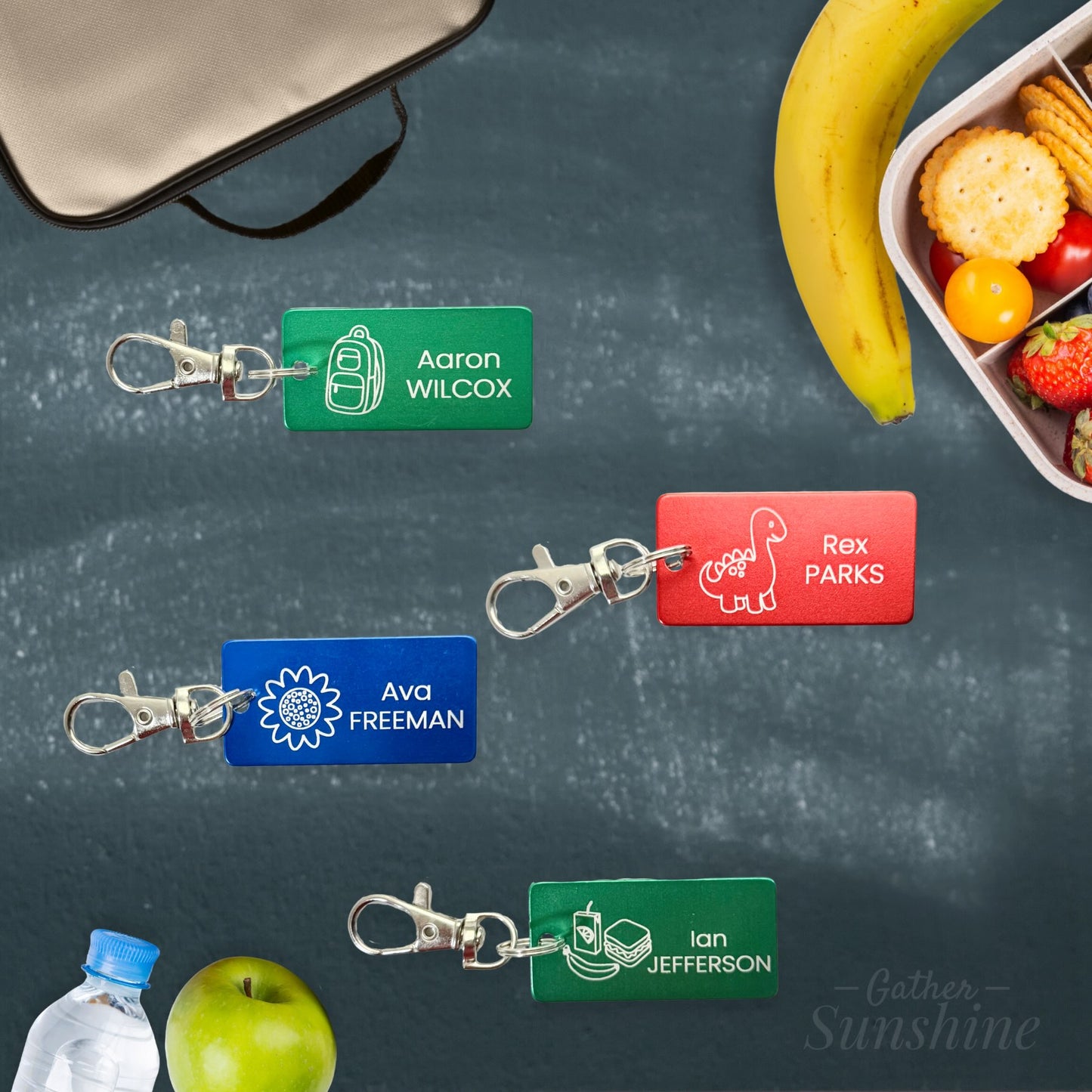 1x2 inch personalized kids school tags with dino, flower, lunch, and backpack icons shown on a chalkboard background. Includes name engraving and lobster clasps