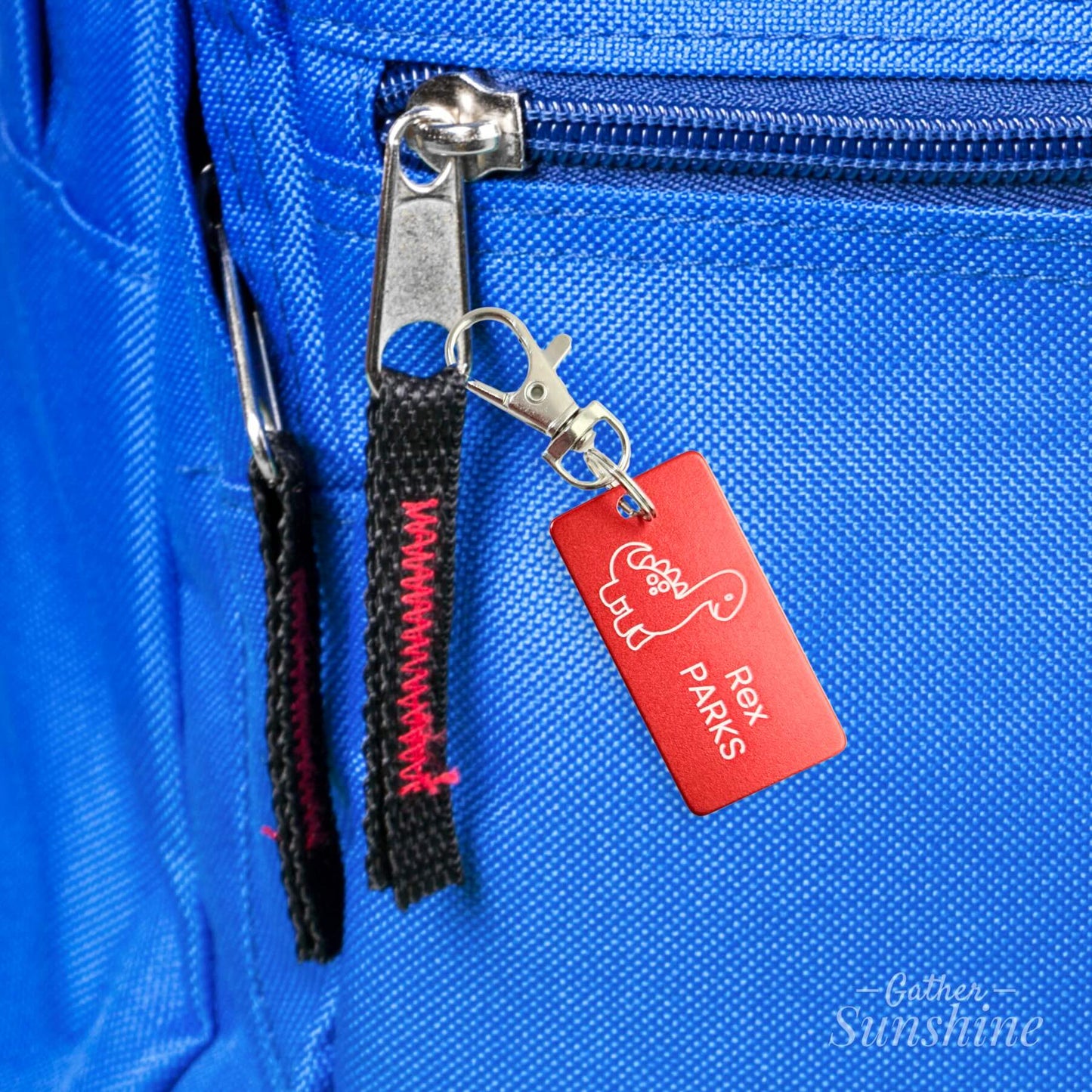 Close-up of red dinosaur kids bag tag clipped to a blue backpack. 1x2 inch tag with lobster clasp and name engraving