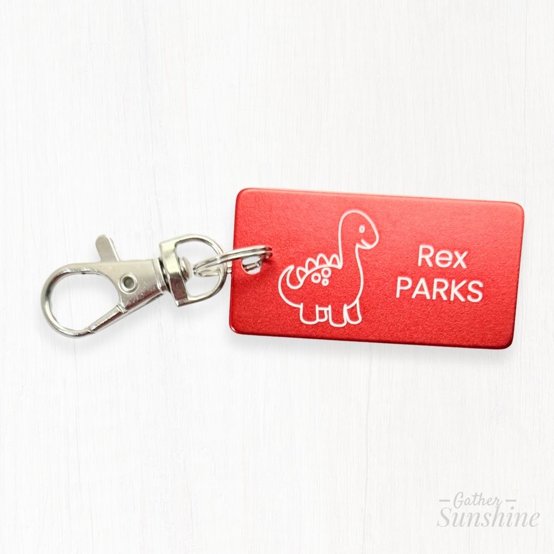 Close-up of red 1x2 inch kids tag with engraved dinosaur design and lobster clasp. Shows detail of personalized engraving
