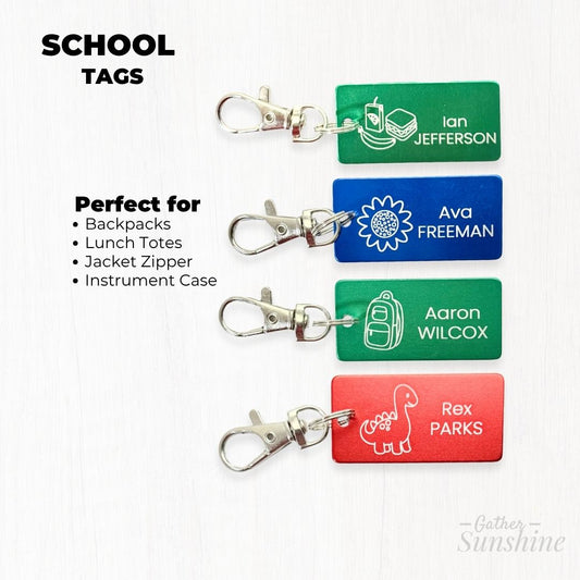 Product photo of 1x2 inch personalized kids school tags in red, green, and blue. Features flower, dino, lunch, and backpack icons with name engraving. Includes overlay text: School Tags – Perfect for backpacks, lunch totes, jacket zippers, and instrument cases