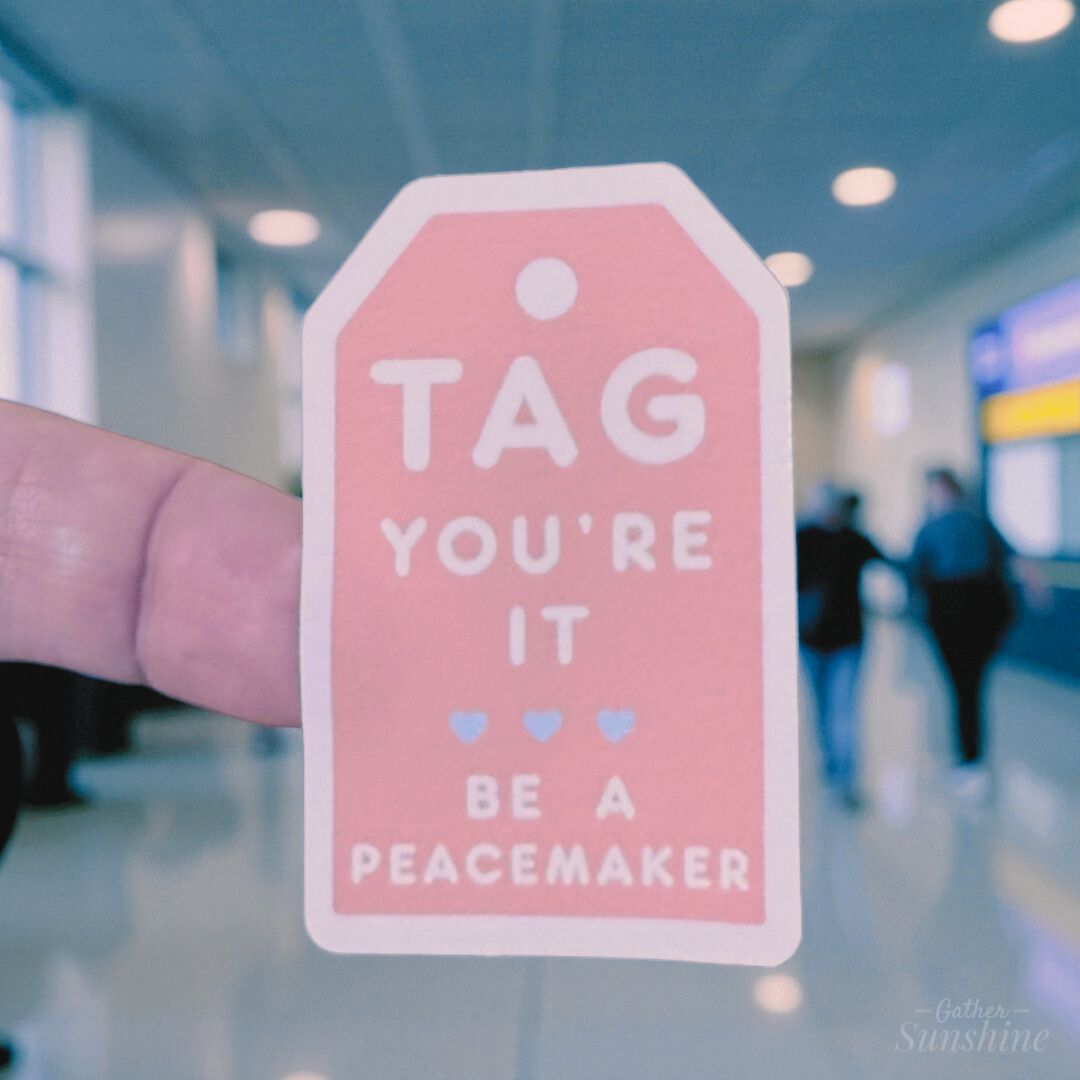 “Tag You’re It – Be a Peacemaker” sticker held in a bright hallway, inspiring acts of peace and positivity – Gathersunshine