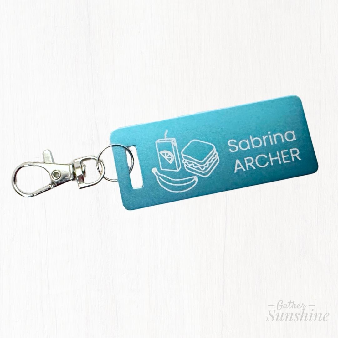 Teal lunchbox tag with illustrations of a drink, sandwich, and banana, and text 'Sabrina Archer' on a white background.