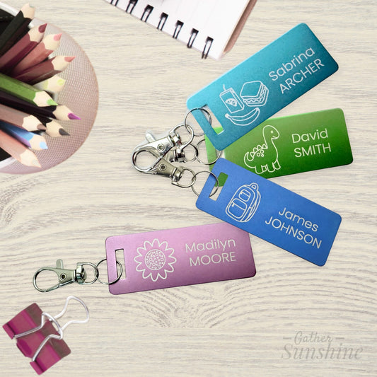 Set of four colorful 1x3 inch kids luggage tags, each with a lobster clasp: a purple flower tag, blue backpack tag, green dinosaur tag, and teal lunch tag — all personalized with names. Displayed on a school desk with colored pencils and paper in the background. Perfect for backpacks, lunch boxes, and more. GatherSunshine.