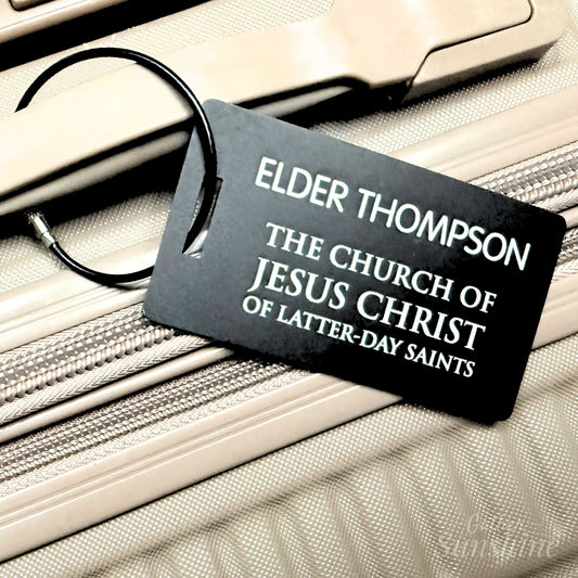 Close-up of engraved missionary luggage tag attached to suitcase zipper, durable black metal tag by Gather Sunshine