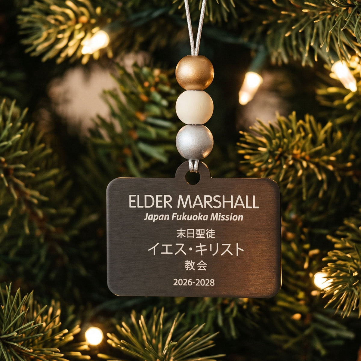 Japanese-language Elder missionary name tag ornament with gold, white, and silver beads hanging among tree branches.