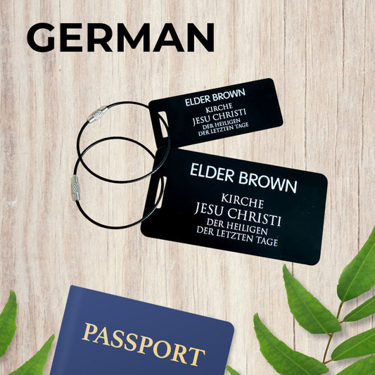 Small (3x1 inch) and large (3.5x2 inch) black luggage tags inspired by LDS missionary name tags in German, on a wooden surface with leaves and a passport by Gather Sunshine.