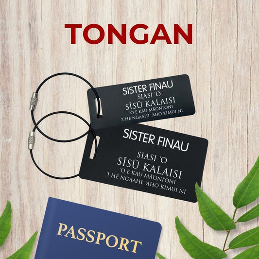 Small (3x1 inch) and large (3.5x2 inch) black luggage tags inspired by LDS missionary name tags, in Tongan with personalized name, on a wooden surface with leaves and a passport by Gather Sunshine.