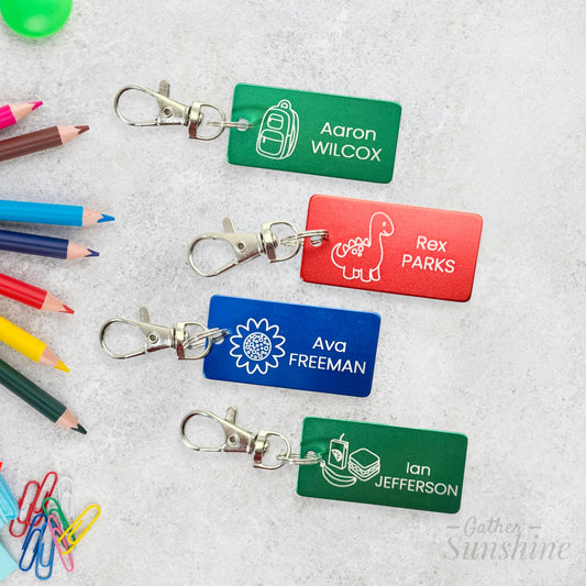 Mockup of 1x2 inch personalized kids school tags with dino, flower, backpack, and lunch icons. Shown on gray background with colored pencils and paperclips on a textured background