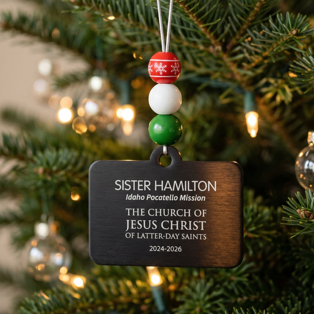 Personalized Sister missionary name tag ornament with red, white, and green beads hanging on a lit Christmas tree.