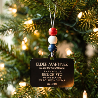 Spanish-language Elder missionary name tag ornament with red, white, and blue beads on a lit Christmas tree.